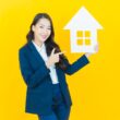 Agent immobilier souriante en costume bleu tenant une maquette blanche de maison et pointant dessus, fond jaune vif.