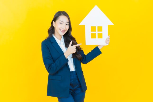 Agent immobilier souriante en costume bleu tenant une maquette blanche de maison et pointant dessus, fond jaune vif.