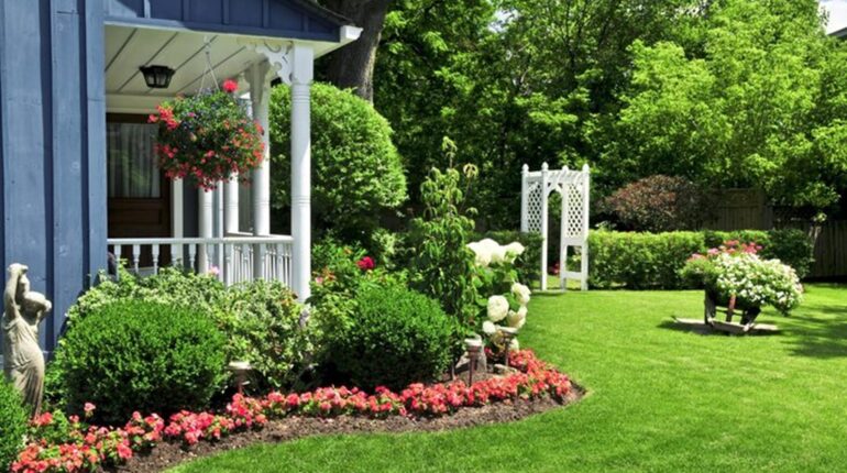 Un jardin bien entretenu avec une pelouse verte, des massifs de fleurs colorées et une maison bleue avec une véranda blanche.