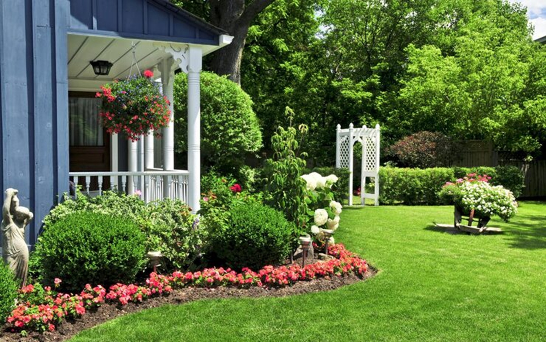 Un jardin bien entretenu avec une pelouse verte, des massifs de fleurs colorées et une maison bleue avec une véranda blanche.