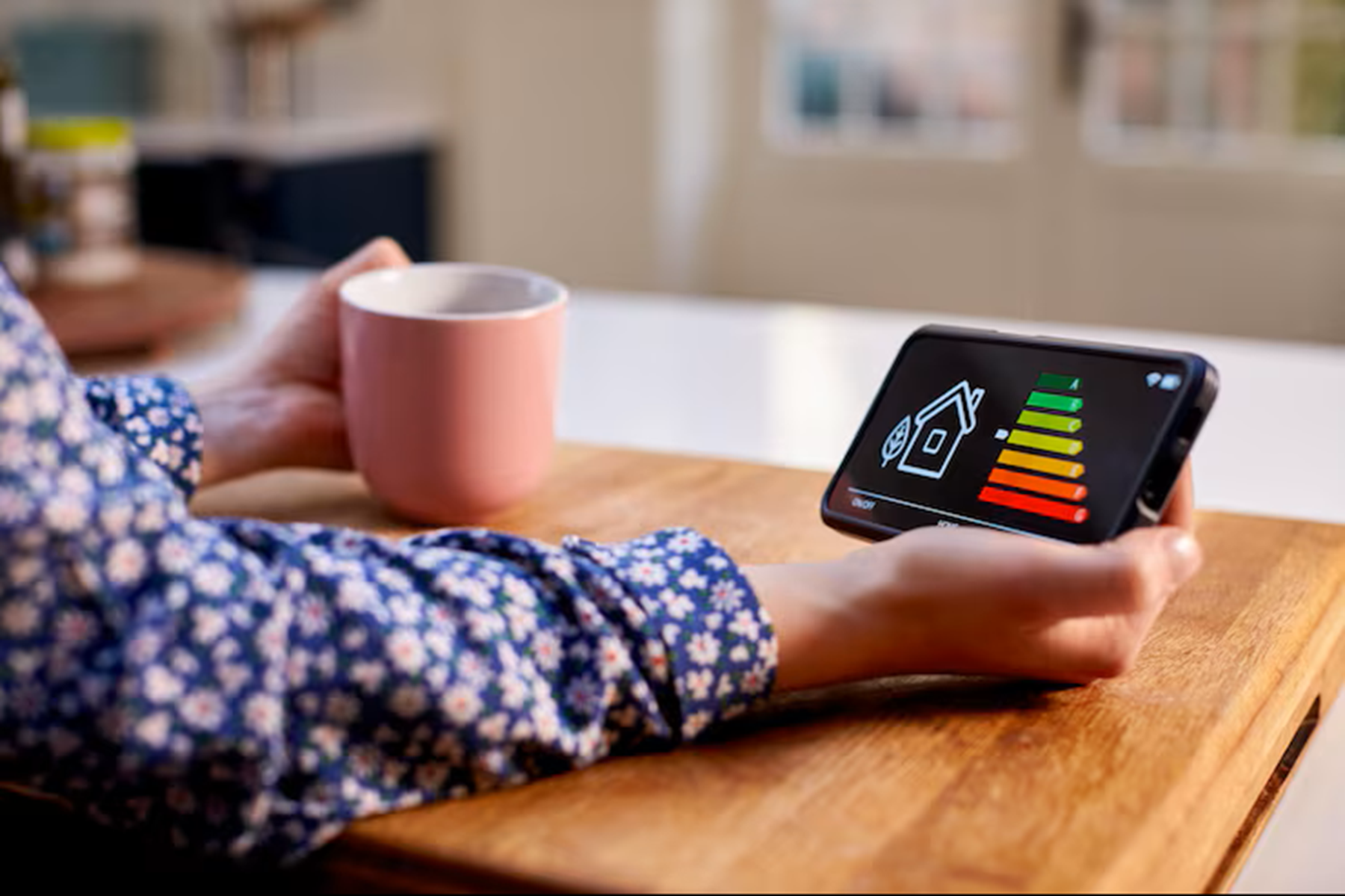 Une personne consulte un diagnostic de performance énergétique (DPE) sur son smartphone tout en tenant une tasse, dans un intérieur lumineux.