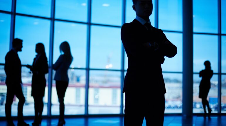 Silhouettes de personnes en costume discutant dans un bureau avec de grandes fenêtres.