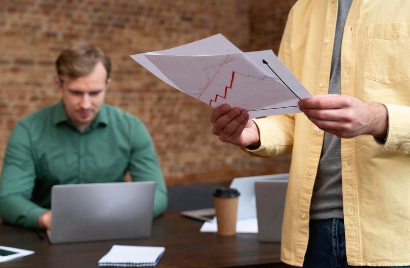 Un homme tient des feuilles avec des graphiques pendant qu’un collègue travaille sur un ordinateur portable.