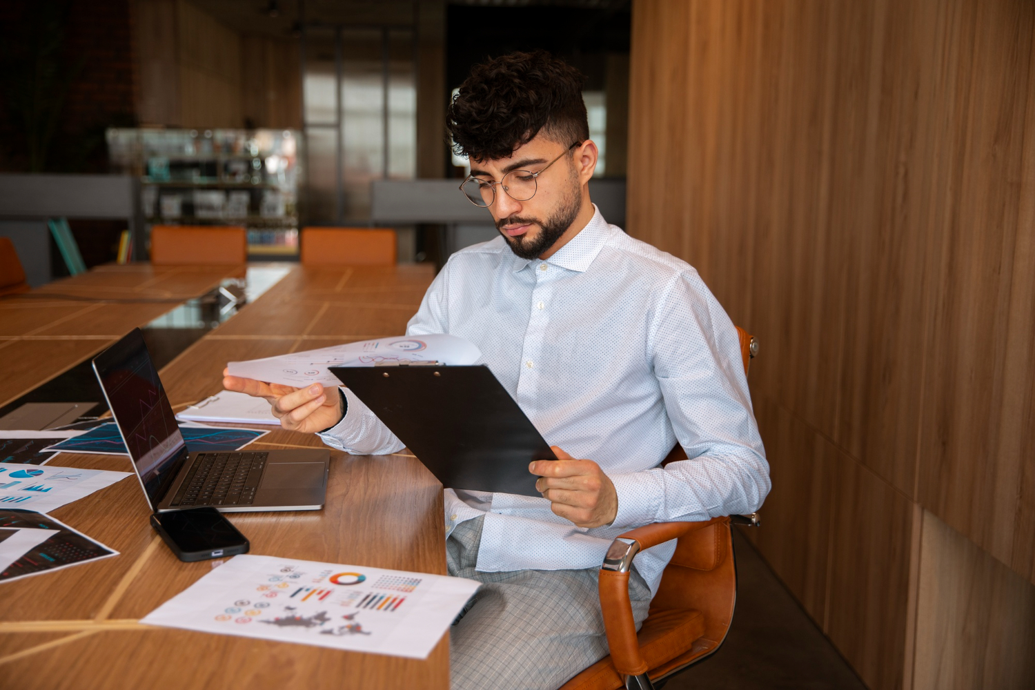 Analyste examinant des rapports sur un porte-bloc avec des graphiques et un ordinateur sur une table de réunion.