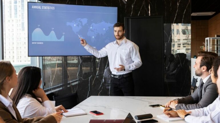 Un présentateur en chemise blanche montre des graphiques statistiques sur un grand écran devant ses collègues assis autour d'une table de réunion.