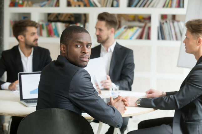 Un homme en costume se retourne pour regarder l'objectif lors d'une réunion d'affaires, entouré de collègues assis autour d'une table avec des ordinateurs et des documents