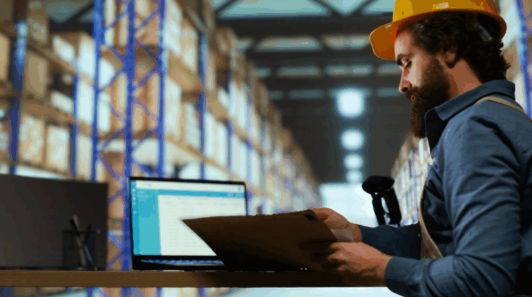 Un homme barbu portant un casque de chantier jaune consulte des documents à un bureau, à côté d'un ordinateur portable ouvert dans un grand entrepôt de logistique